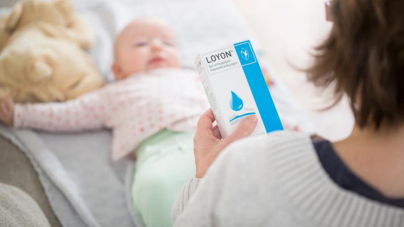 Baby liegt vor der Mutter auf einer Decke, während die Mutter eine Packung LOYON® in der Hand hält und diese liest.